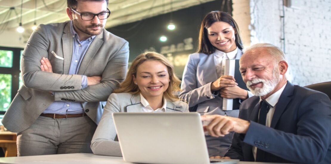 A diverse group of four business people gathered around a table for a meeting looking at a laptop and reviewing Business Central & Teams Integration