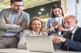 A diverse group of four business people gathered around a table for a meeting looking at a laptop and reviewing Business Central & Teams Integration